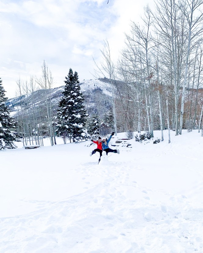 two blonde girls in ski gear jumping the snow with a snowman