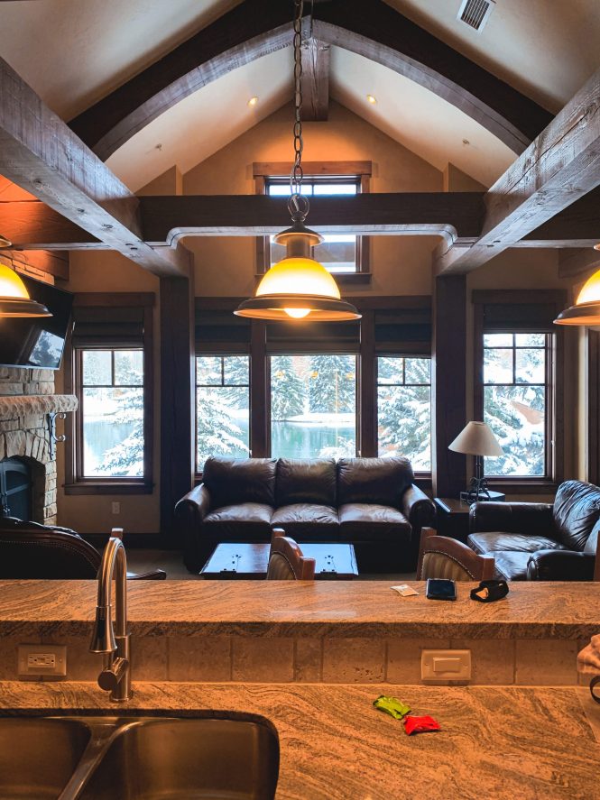 winter cabin kitchen overlooking living area with snow and tall pine trees in the window view
